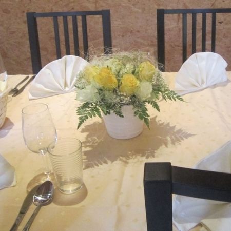 Runder Tisch mit gelbem Blumengesteck in weißem Topf, Gläsern und weiß gefalteten Servietten.
