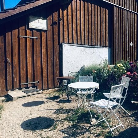 Gemütliche Sitzgruppe mit weißen Metallstühlen und Tisch vor einem Holzhaus mit Blumengarten.