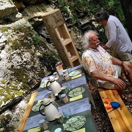 Gedeckter Holztisch mit Frühstücksgeschirr im Freien an einer moosbewachsenen Felswand, zwei Personen sitzen bzw. stehen daneben.