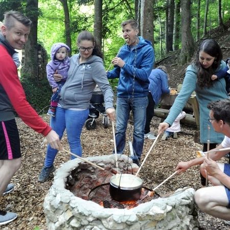 Gruppe von Erwachsenen und Kindern röstet Brotstücke an langen Stöcken über einem offenen Feuer mit Fondue-Topf im Wald.